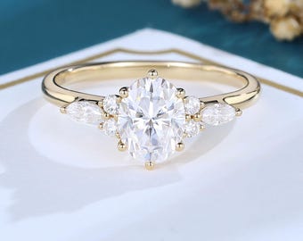 May include: A gold ring featuring a large oval-cut center stone held by six prongs. The center stone is flanked by two smaller round stones on each side and one marquise-cut stone on the far left and right, all set on a polished gold band.
