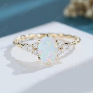 May include: A gold ring featuring a central oval-shaped iridescent opal stone flanked by two marquise-cut clear stones and two smaller round clear stones on each side. The band has a twisted rope-like texture, creating a detailed and elegant appearance.
