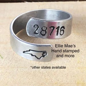 May include: A silver ring with the state of North Carolina and the number 28716 stamped on it. The ring is made by Ellie Mae's Hand Stamped and more. Other states are available.