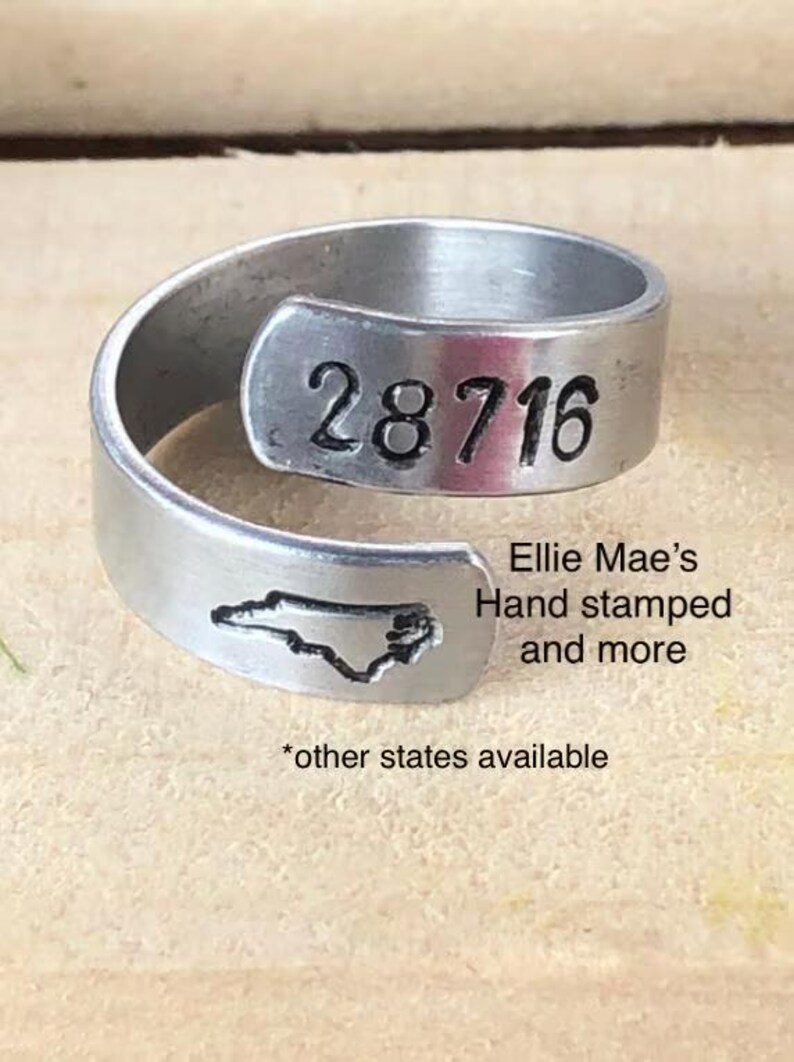 May include: A silver ring with the state of North Carolina and the number 28716 stamped on it. The ring is made by Ellie Mae's Hand Stamped and more. Other states are available.