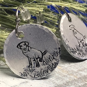 K&ouml;nnte beinhalten: Ein Paar silberne Ohrringe mit einem Hundesilhouetten-Design. Jeder Ohrring ist eine runde Metallscheibe mit einer detaillierten Umrandung eines Hundes, der im Gras steht. Die Ohrringe haben einen einfachen Hakenverschluss.