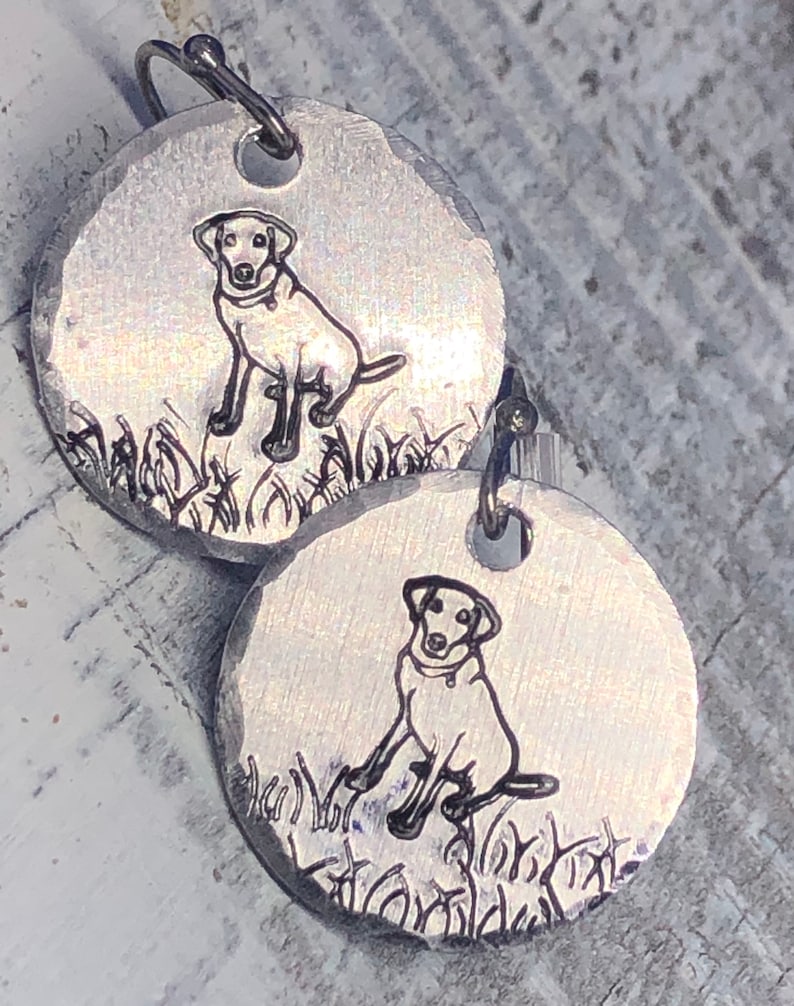 K&ouml;nnte beinhalten: Ein Paar silberne Ohrringe mit einem Schwarzwei&szlig;bild eines Hundes, der im Gras sitzt. Die Ohrringe sind rund und haben eine geh&auml;mmerte Textur.