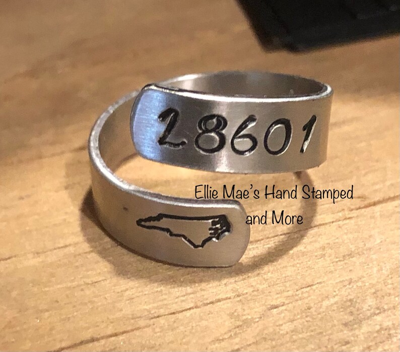 May include: A silver ring with the number "18601" engraved on it. The ring also features a silhouette of the state of North Carolina. The ring is made by Ellie Mae's Hand Stamped and More.