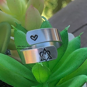 May include: A silver-colored wrap ring with etched designs. One end features a heart, the other a heart intertwined with a triangle. The ring is displayed on a bed of green succulent leaves. The text "Ellie Mae's Hand stamped and more" is visible.