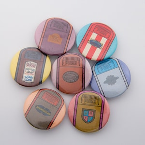 May include: Seven round, colorful buttons with a cartoon image of a trash can on each. The trash cans have different colors and designs, including a red and white striped can, a brown can with a gold trim, and a blue can with a white cloud.