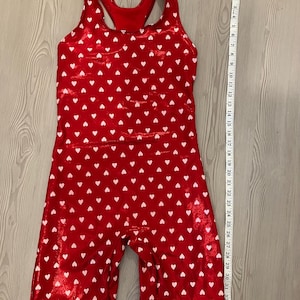 May include: Red jumpsuit with white heart pattern. The sleeveless one-piece garment has a scoop back and short legs. A measuring tape shows the jumpsuit is approximately 34 inches long.