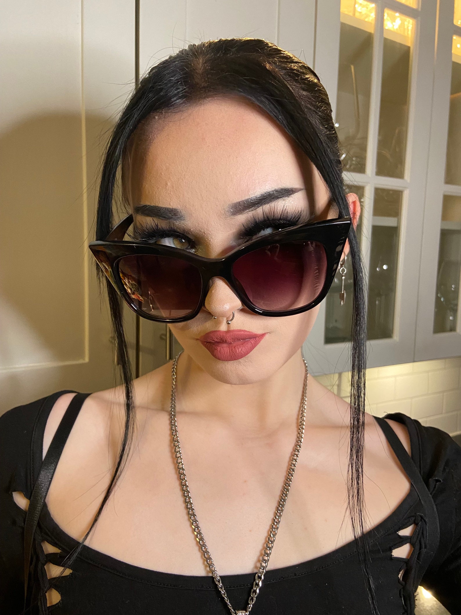 Close-up of gothic woman wearing oversized black cat-eye sunglasses, bold lashes, silver septum ring, and a black cutout top