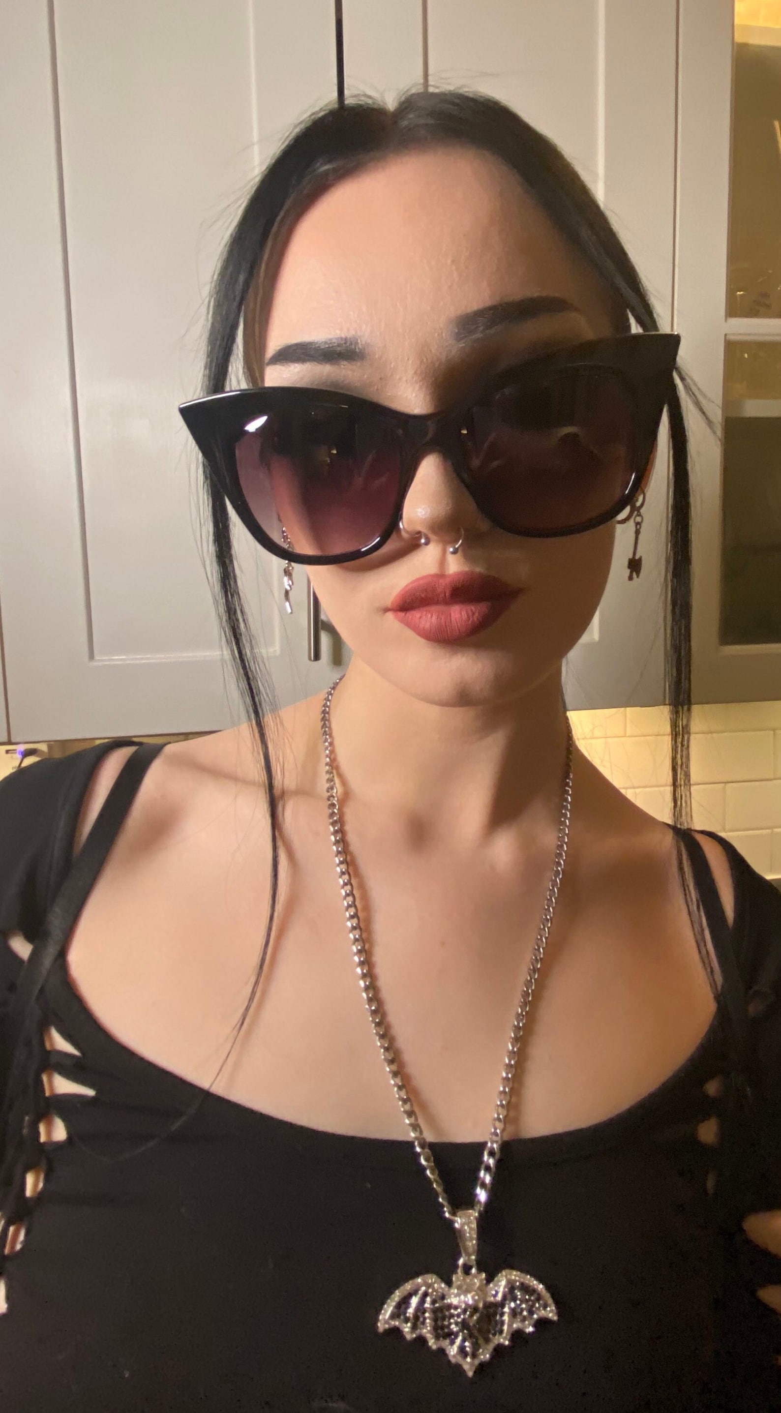 Gothic woman in black cat-eye sunglasses, bat pendant necklace, and lace-up black top with silver septum ring and earrings