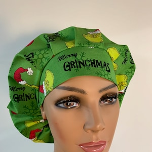 May include: Green scrub cap featuring a Grinch-themed pattern. The cap has a band with the text "Merry Grinchmas" in black. The design includes images of the Grinch, snowflakes, and other Christmas-themed elements. The cap is made of a soft, likely cotton, fabric.