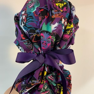 May include: A purple scrub cap featuring a repeating graphic print of various animated villain characters in vibrant colors. The cap has a gathered back design and is secured with a solid dark purple grosgrain ribbon tied into a bow.