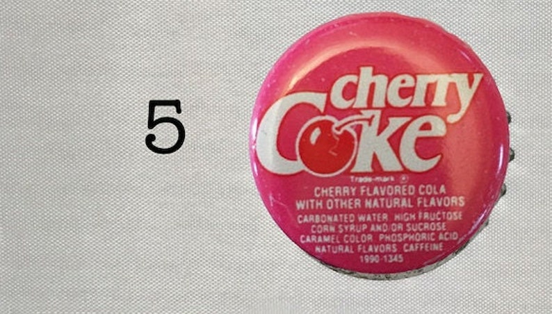 Może przedstawiać: R&oacute;żowa zakrętka butelki z białym tekstem, kt&oacute;ry brzmi "cherry Coke" i zawiera grafikę wiśni. Tekst pod logo brzmi "Cherry Flavored Cola with Other Natural Flavors" i zawiera składniki i daty.