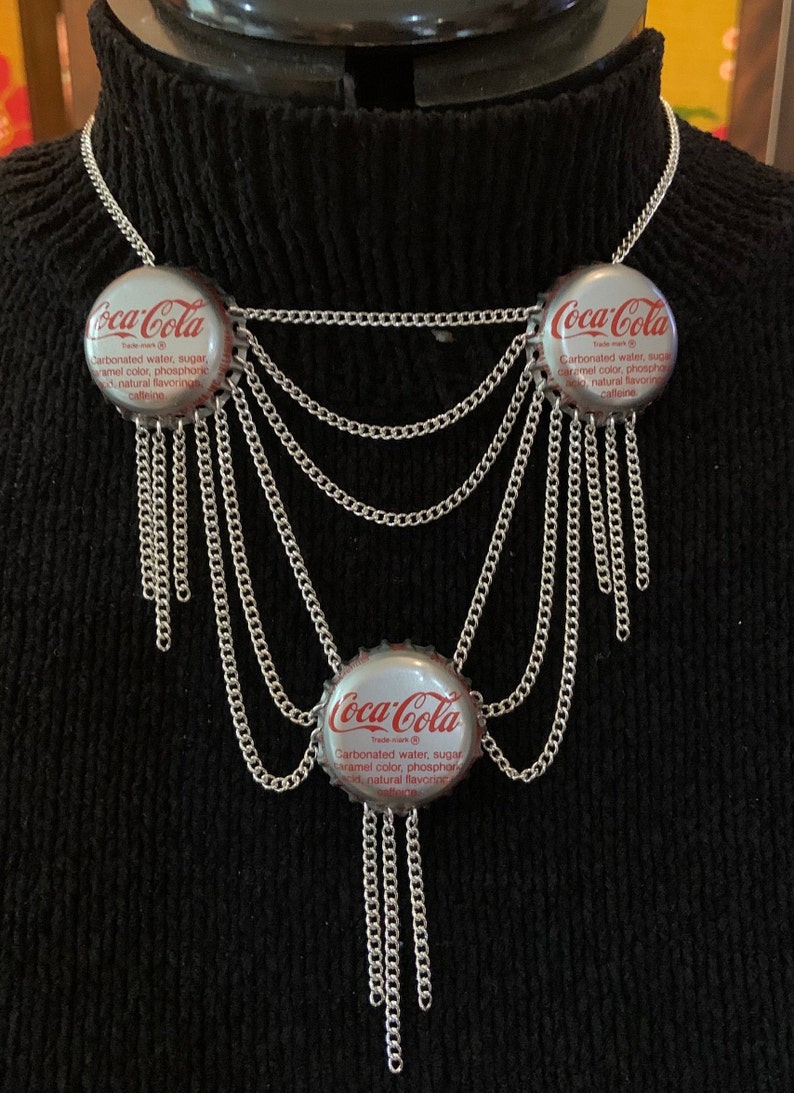 Może przedstawiać: Srebrny naszyjnik z łańcuszkiem z trzema korkami od butelek Coca-Cola jako zawieszkami. Na korkach widnieje logo Coca-Cola i tekst: "Woda gazowana, cukier, barwnik karmelowy, kwas fosforowy, naturalne aromaty, kofeina".