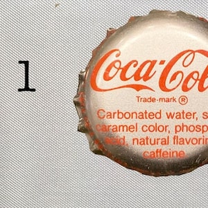 Może przedstawiać: Srebrna zakrętka butelki z logo Coca-Cola i tekstem "Trade-mark R Woda gazowana, cukier, barwnik karmelowy, kwas fosforowy, naturalne aromaty, kofeina".