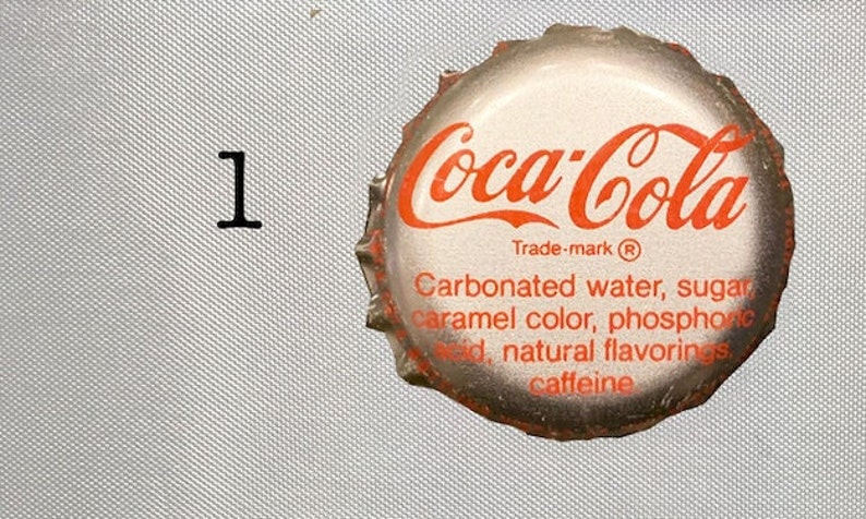 Może przedstawiać: Srebrna zakrętka butelki z logo Coca-Cola i tekstem "Trade-mark R Woda gazowana, cukier, barwnik karmelowy, kwas fosforowy, naturalne aromaty, kofeina".