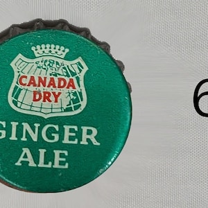 Może przedstawiać: Zielona zakrętka butelki z białym logo tarczy, na kt&oacute;rym widnieje napis "Canada Dry" i "Ginger Ale".