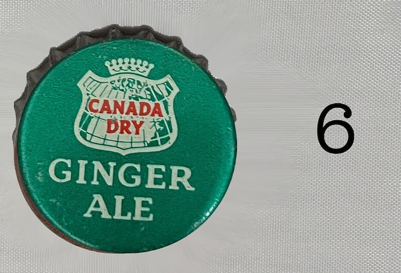 Może przedstawiać: Zielona zakrętka butelki z białym logo tarczy, na kt&oacute;rym widnieje napis "Canada Dry" i "Ginger Ale".