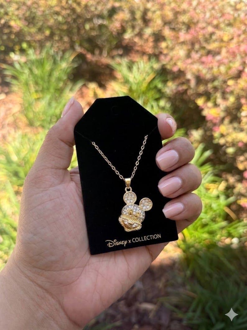 May include: Gold-coloured necklace with a Mickey Mouse pendant, set with clear crystals. The pendant hangs from a fine chain and is presented on a black card. The card has the text "Disney x Collection".