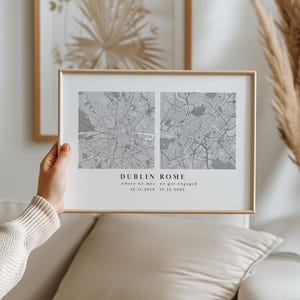 May include: A gold-framed print displaying two grey city maps, one of Dublin and one of Rome. The text "DUBLIN ROME" is centered beneath the maps, with dates below marking important occasions. The print is held up.