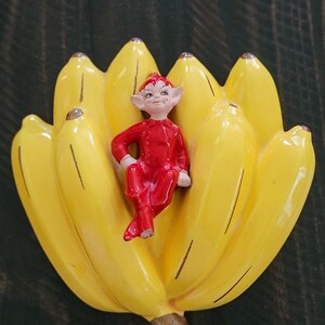May include: A ceramic figurine of a red-clad elf sitting on a bunch of yellow bananas.