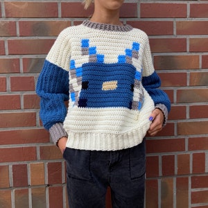 Puede incluir: Un su&eacute;ter de crochet blanco con un dise&ntilde;o pixelado azul y gris en la parte delantera. El su&eacute;ter tiene mangas largas y cuello redondo.