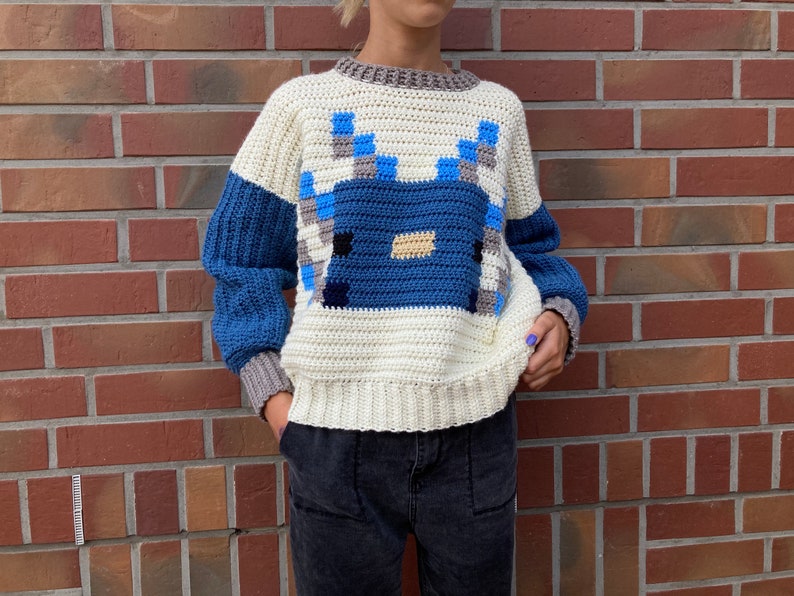 Puede incluir: Un su&eacute;ter de crochet blanco con un dise&ntilde;o pixelado azul y gris en la parte delantera. El su&eacute;ter tiene mangas largas y cuello redondo.
