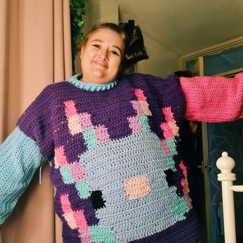 Puede incluir: Un su&eacute;ter de crochet morado, azul, rosa y verde con un dise&ntilde;o pixelado de un animal de dibujos animados. El su&eacute;ter tiene mangas largas y cuello redondo.