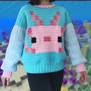 Puede incluir: Un su&eacute;ter de crochet azul turquesa con un dise&ntilde;o de ajolote rosa y blanco en la parte delantera. El su&eacute;ter tiene mangas largas y cuello redondo.