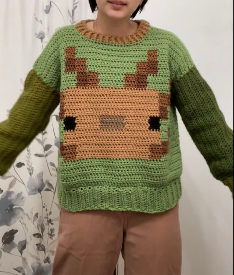 Puede incluir: Su&eacute;ter de crochet verde con un dise&ntilde;o de ciervo marr&oacute;n. El su&eacute;ter tiene mangas largas y cuello redondo.