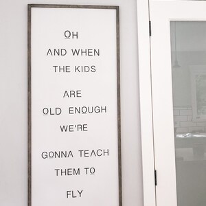 May include: A rectangular wooden sign with a white background and dark gray frame. The sign features black text that reads "Oh and when the kids are old enough we're gonna teach them to fly." A black arrow design is at the bottom.