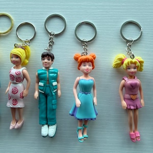 Puede incluir: Una colección de llaveros de muñecas en miniatura. Las muñecas visten varios atuendos, incluyendo vestidos y pantalones. Cada llavero tiene un anillo plateado para sujetar a llaves o bolsos.