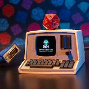 Puede incluir: Un miniordenador retro con una pantalla que muestra "D64" y un logotipo geométrico. Un dado de veinte caras rojo y dorado está encima del ordenador. Un cable VGA azul está conectado. La pantalla dice "Press Any Key" y "Double Click for All Color".
