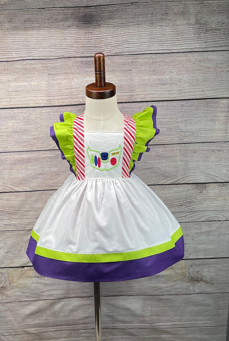 May include: White dress with green and purple trim and a red and white striped bodice. The dress has an embroidered image of Buzz Lightyear's face with the text "SPACE RANGER".