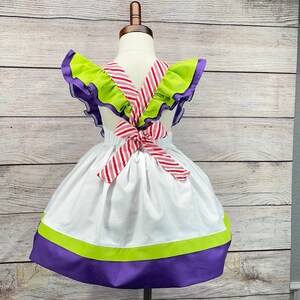 May include: White dress with green and purple trim and a red and white striped bow in the back.