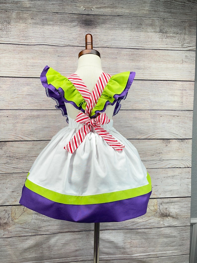 May include: White dress with green and purple trim and a red and white striped bow in the back.