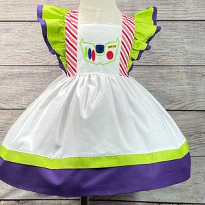 May include: White dress with green and purple trim. The dress has a red and white striped bodice with a green ruffle on the shoulders. The front of the dress has an embroidered image of Buzz Lightyear from the Toy Story movie.