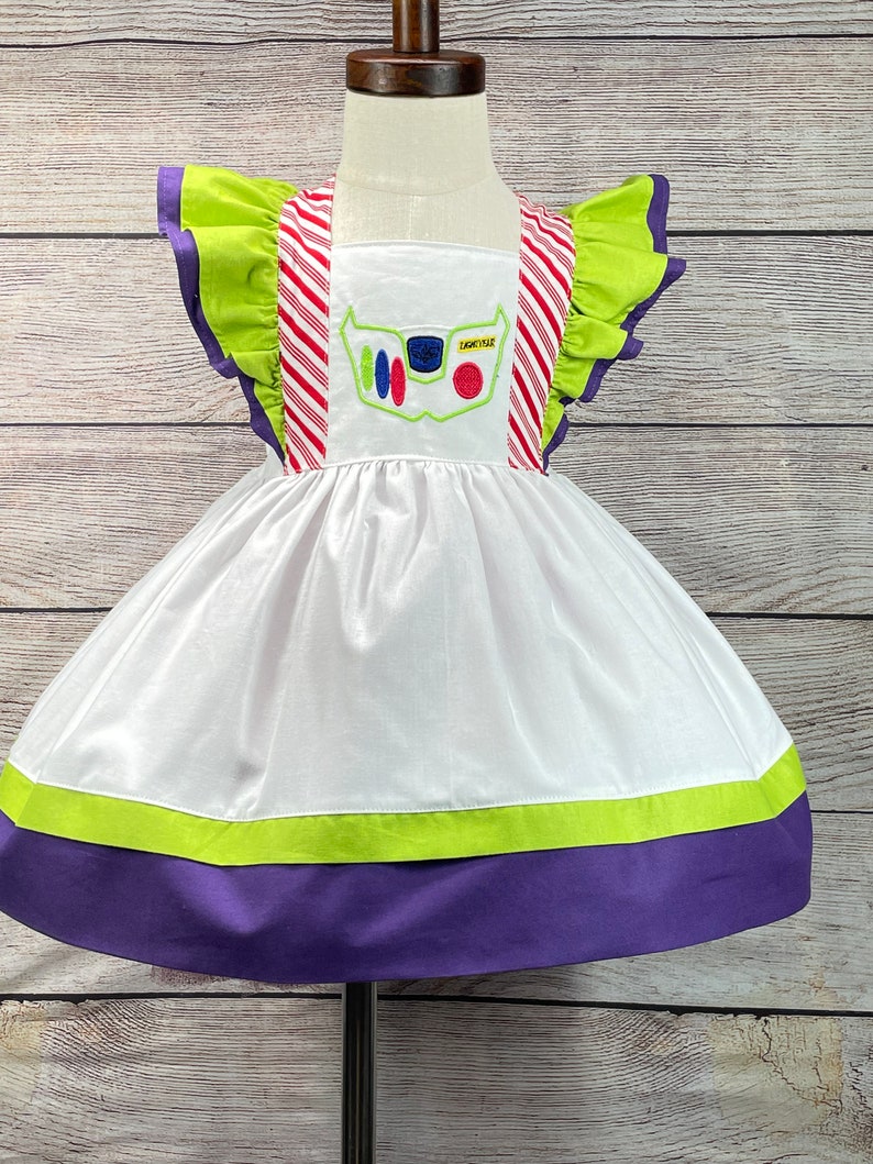 May include: White dress with green and purple trim. The dress has a red and white striped bodice with a green ruffle on the shoulders. The front of the dress has an embroidered image of Buzz Lightyear from the Toy Story movie.