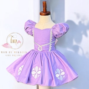 May include: A purple dress with white embroidered flowers and a white lace trim. The dress has puffed sleeves and a sweetheart neckline. "Mom by Mikaela Girl Dresses" is written on the bottom left corner of the image.