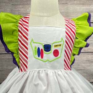 May include: White dress with green and red striped straps and a green ruffle on the sleeves. The dress has an embroidered image of Buzz Lightyear's chest panel with the text "Lightyear" on it.
