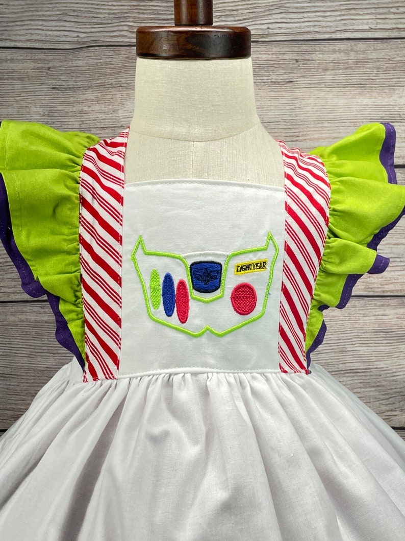 May include: White dress with green and red striped straps and a green ruffle on the sleeves. The dress has an embroidered image of Buzz Lightyear's chest panel with the text "Lightyear" on it.