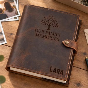 May include: A dark brown leather photo album with a strap and brass button closure. The cover features an engraved tree icon, the text "OUR FAMILY MEMORIES," and the name "LARA" at the bottom. The album has visible stitching along the edges.