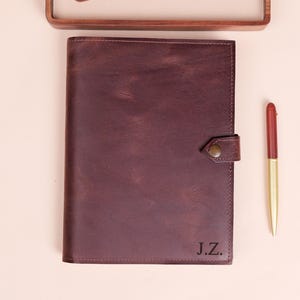 May include: A dark burgundy leather notebook cover with a snap closure and the initials J.Z. embossed on the bottom right corner. The notebook is accompanied by a gold and red pen, a green leather card holder, and a wooden tray.