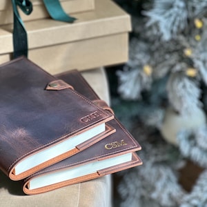 May include: Two brown leather journals with gold embossed initials "CRH" and "CSM" on the covers. The journals are stacked on top of each other and are sitting on a brown surface.