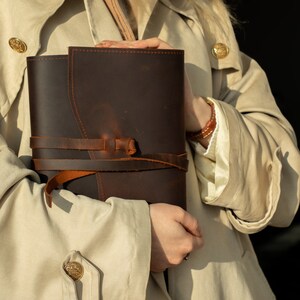 May include: A brown leather journal with a strap closure. The journal is held by a person wearing a beige trench coat.