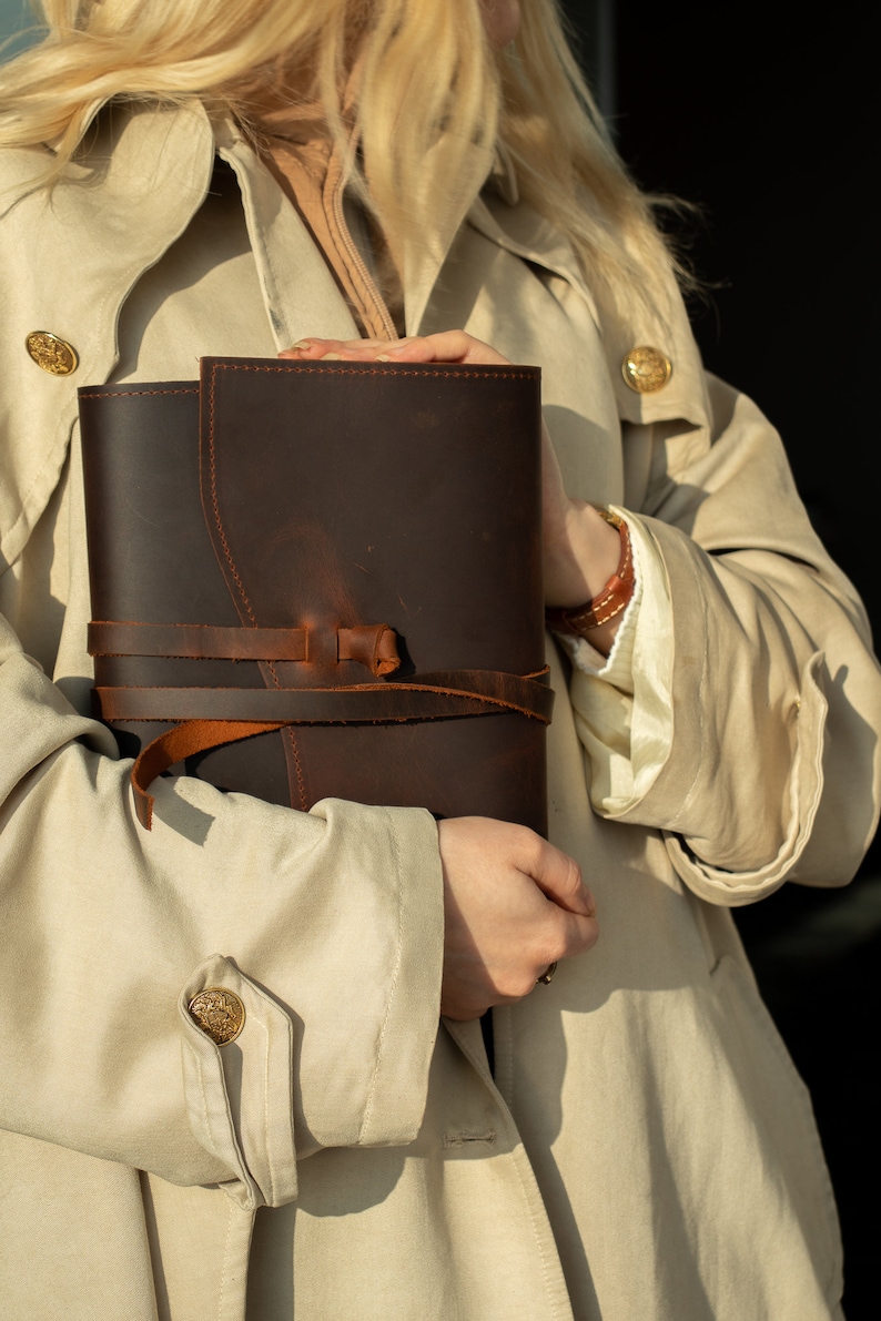 May include: A brown leather journal with a strap closure. The journal is held by a person wearing a beige trench coat.