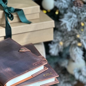 May include: Two brown leather journals with gold embossed initials "CRH" and "CSM" on the covers. The journals are stacked on top of each other and are in focus. The background is out of focus and shows a Christmas tree with white ornaments.
