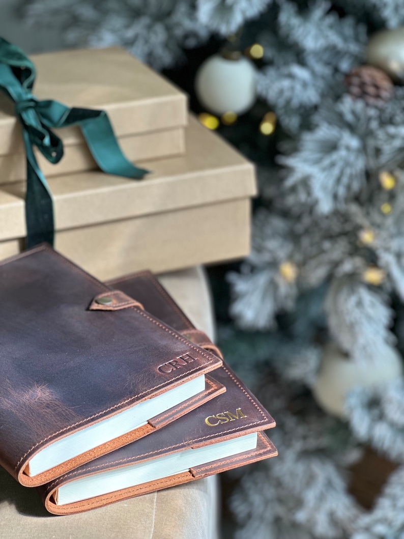 May include: Two brown leather journals with gold embossed initials "CRH" and "CSM" on the covers. The journals are stacked on top of each other and are in focus. The background is out of focus and shows a Christmas tree with white ornaments.