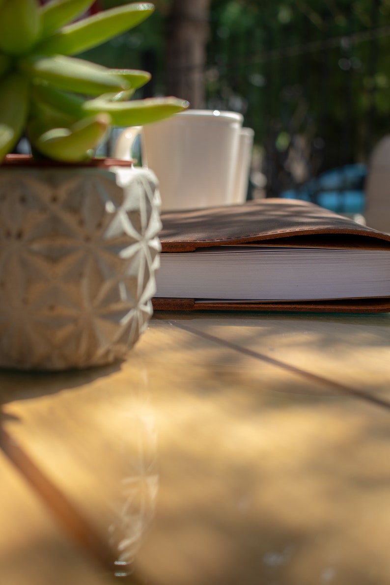May include: A brown leather-bound journal with white pages sits on a wooden table next to a white ceramic planter with a succulent plant.