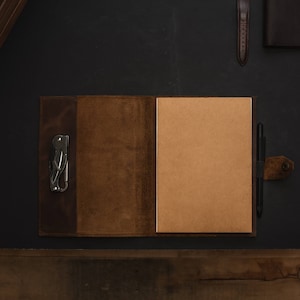 May include: A brown leather journal with a pen and a multi-tool attached to the cover. The journal is open to a blank page.