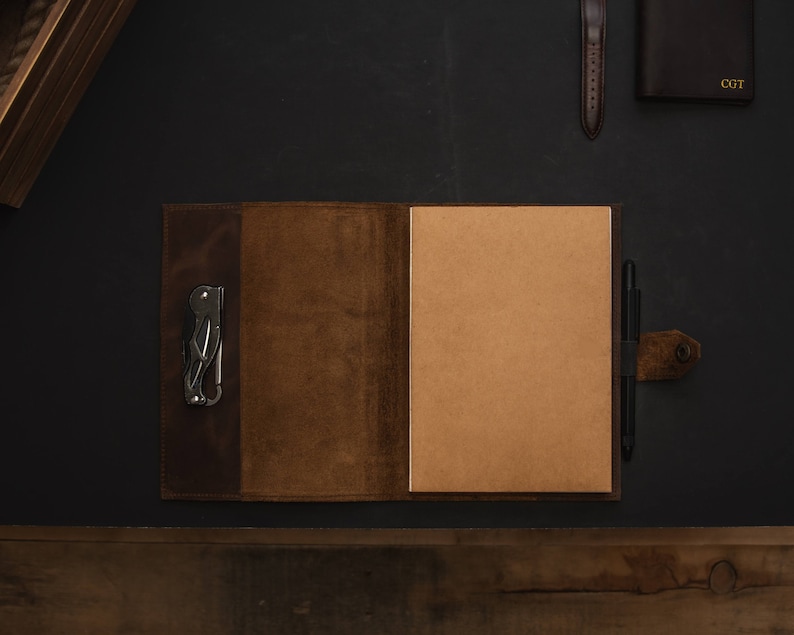 May include: A brown leather journal with a pen and a multi-tool attached to the cover. The journal is open to a blank page.