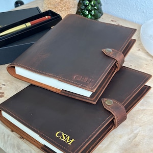 May include: Two brown leather journals with personalized initials, "CSM" and "GRH", in gold lettering. The journals have a flap closure with a metal button.